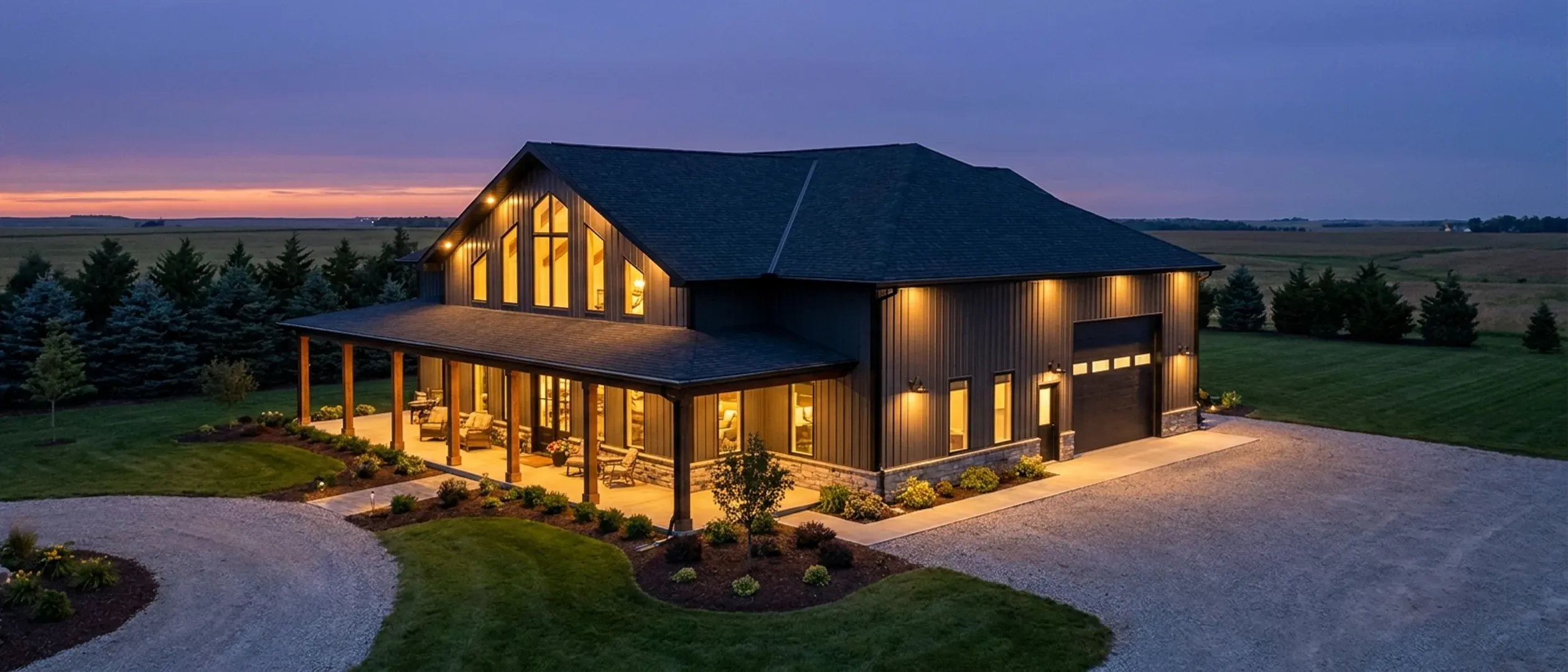 A barndominium at dusk with warm light glowing from every window and exterior lighting along the porch and roofline, a dark windbreak and open farmland behind it.