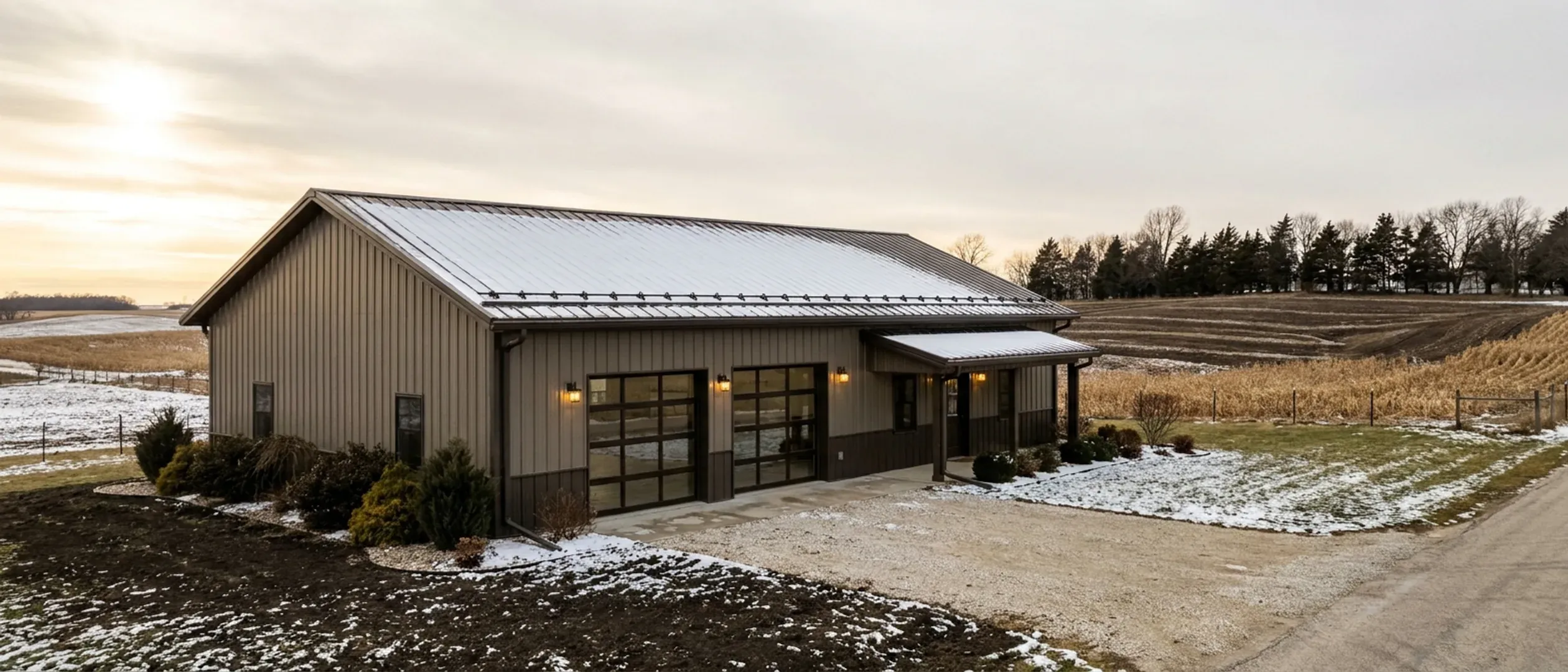 A completed barndominium in late winter viewed from a low angle, showing its snow-dusted standing seam roof, vertical wall panels, gutters and exterior wall lights.
