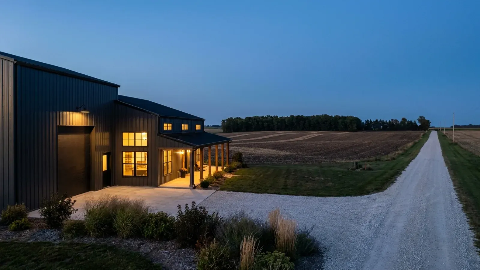 A finished barndominium at dusk with warm interior lighting glowing through its large windows.