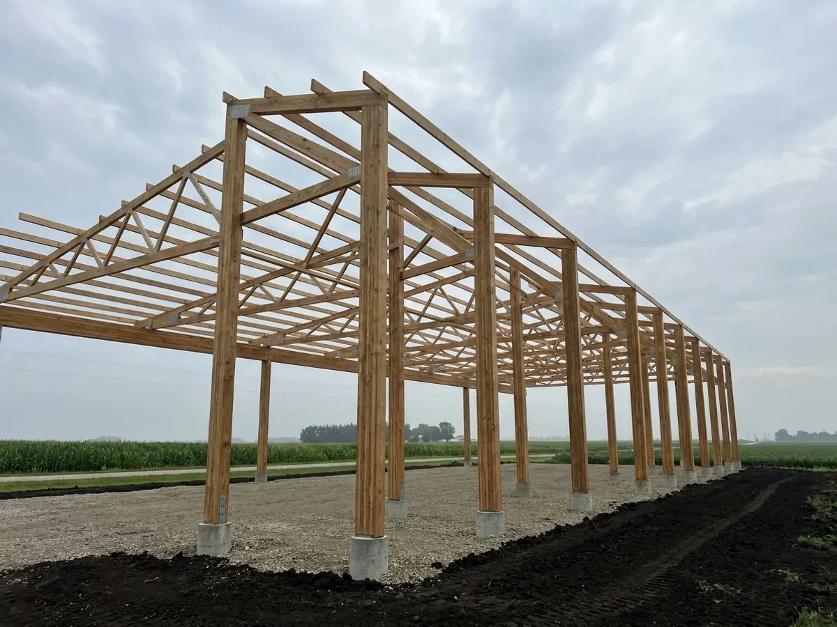 A post-frame home under construction, showing laminated timber columns on concrete piers and wide-span roof trusses with no interior bearing walls.